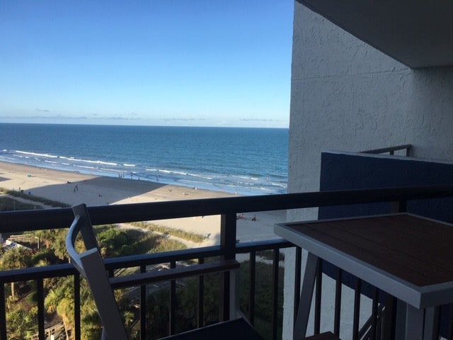 A balcony view overlooking the Atlantic Ocean displays a wide expanse of sandy beach and rhythmic waves. The horizon meets a clear blue sky, while a small outdoor table and chairs are positioned near the railing, inviting relaxation amidst the scenic vista.