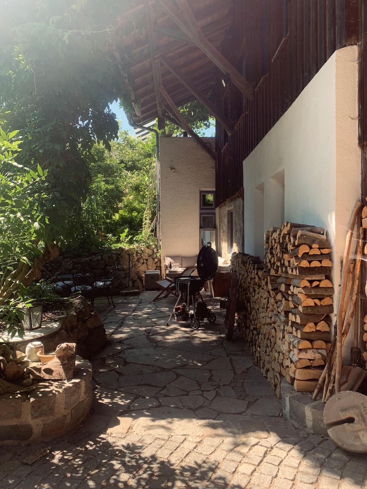 A stone-paved outdoor area is enhanced by greenery and a rustic wooden structure. A small table and chairs are positioned in the shade, providing a cozy setting. Stacked firewood lines the wall, and a grill is set up nearby, inviting outdoor cooking experiences.