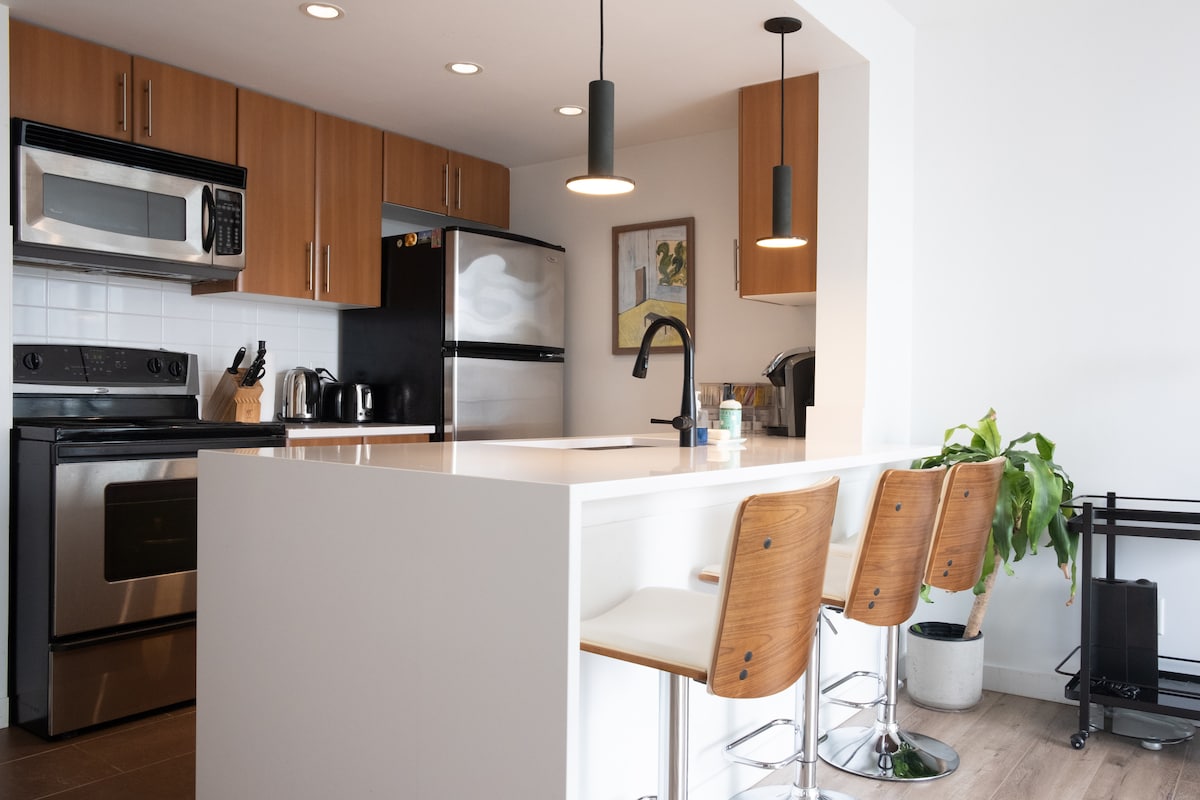 The kitchen features modern cabinetry and stainless steel appliances, including an oven and microwave. A large island with seating for three is highlighted, alongside a potted plant and framed artwork on the wall, enhancing the functional yet stylish environment.