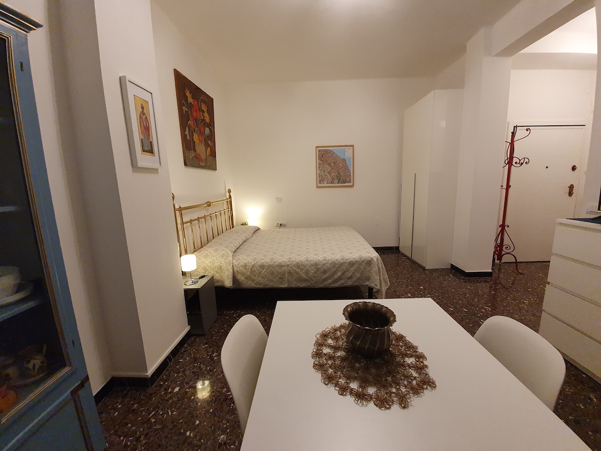 A bright, modern studio apartment features a spacious bed with neutral bedding. A dining table with white chairs occupies the foreground. Artwork decorates the walls, including a framed landscape piece. Soft lighting enhances the clean, inviting space.