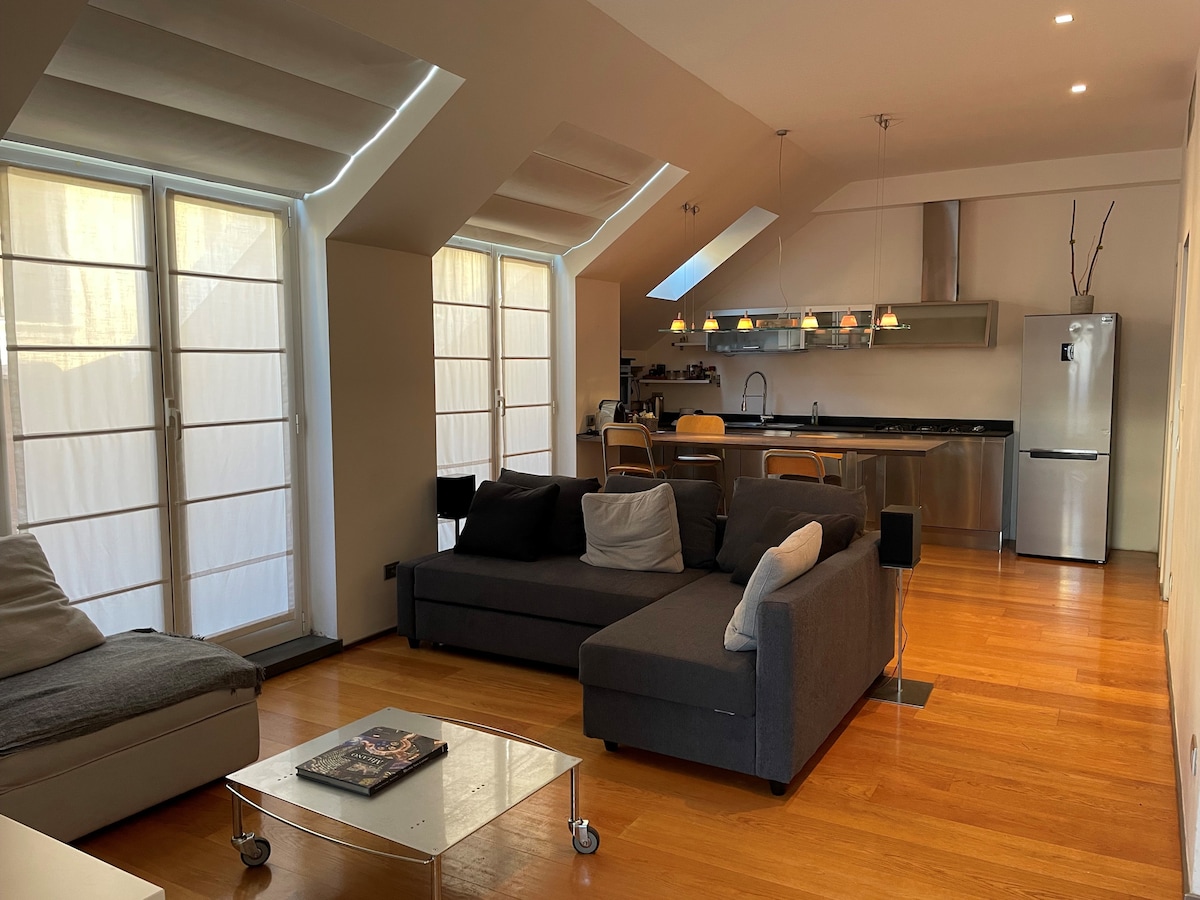 A spacious living area features large windows allowing ample natural light. A sectional grey sofa with contrasting cushions is positioned in front of a glass coffee table. A modern kitchen can be seen in the background, highlighted by sleek appliances and pendant lighting.