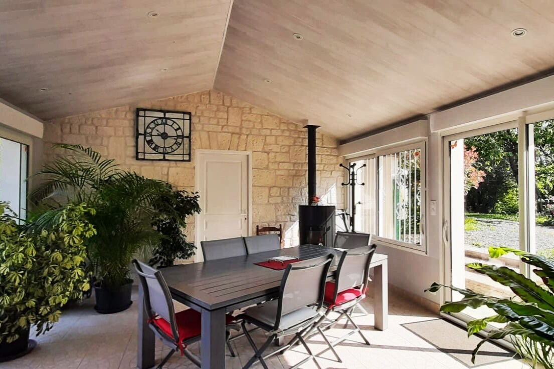 An indoor dining area features a large table surrounded by gray chairs with red cushions. A wood stove adds warmth, while large windows provide a view of the outdoors. Potted plants are positioned throughout, contributing to a serene atmosphere.