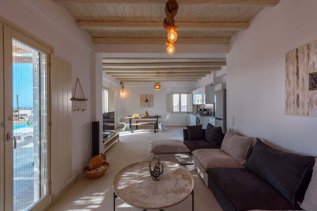 The open-plan living area features a cozy sofa arrangement and a stylish coffee table. Natural light floods the room through large windows, offering views of the outdoor space. Exposed wooden beams and decorative elements create a welcoming ambiance.