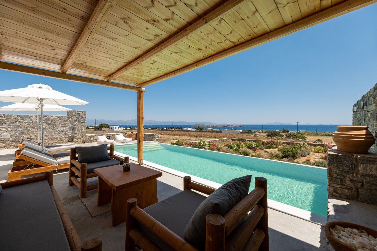 A shaded outdoor seating area features wooden furniture, including a coffee table and comfortable sofas. The space offers direct views of the swimming pool and distant Aegean Sea. An umbrella provides shade, while vibrant greenery and a stone wall outline the quiet surroundings.