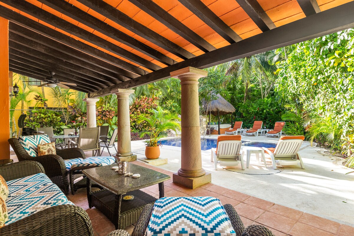 A shaded outdoor area features comfortable lounge chairs and a table set on tiled flooring. Lush greenery surrounds a private pool, with a thatched-roof cabana visible in the background. The ambiance is enhanced by well-placed columns and vibrant decor.