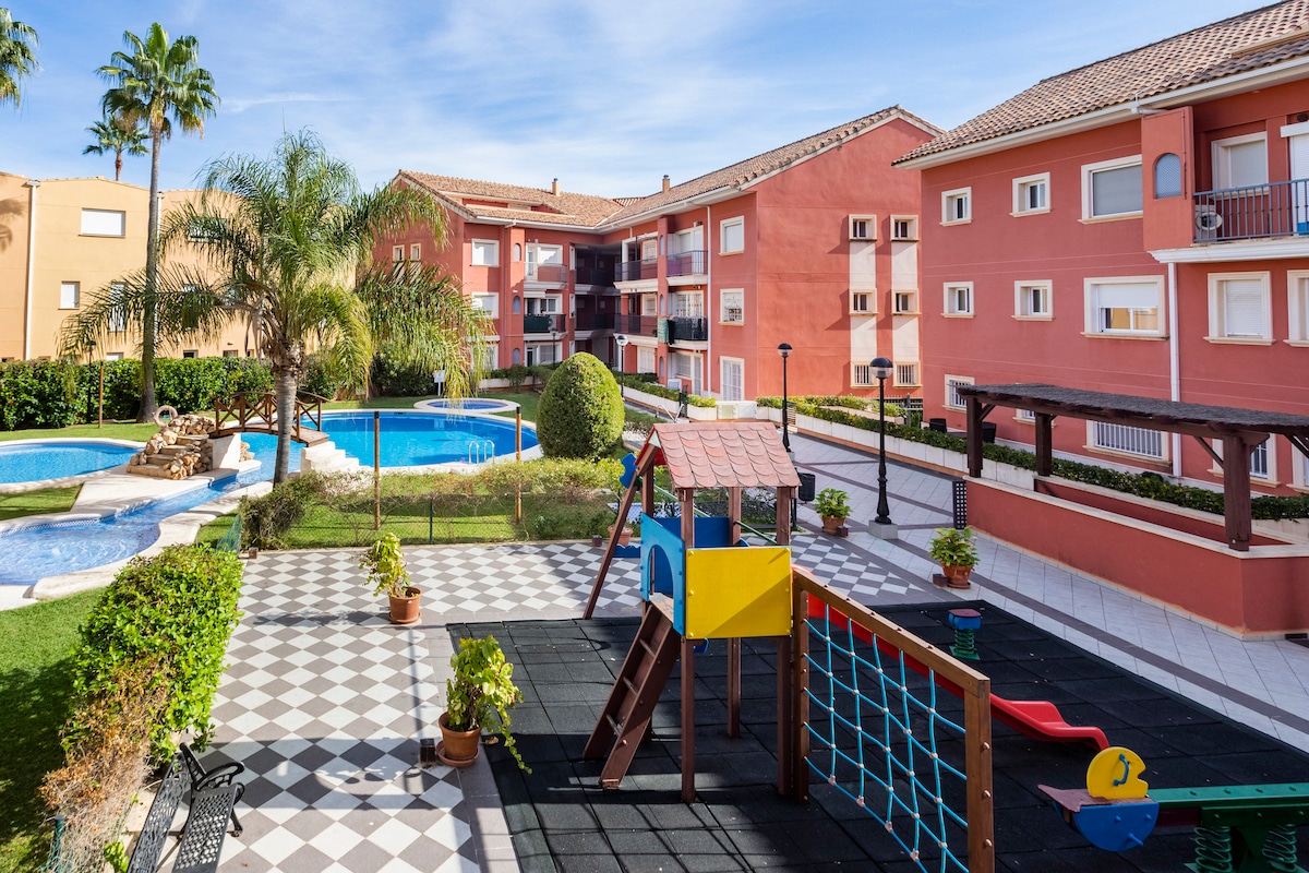 A community area features a playground with a slide and climbing frame, surrounded by well-maintained grass and decorative plants. The view includes a swimming pool and residential buildings, highlighting a serene environment for families.