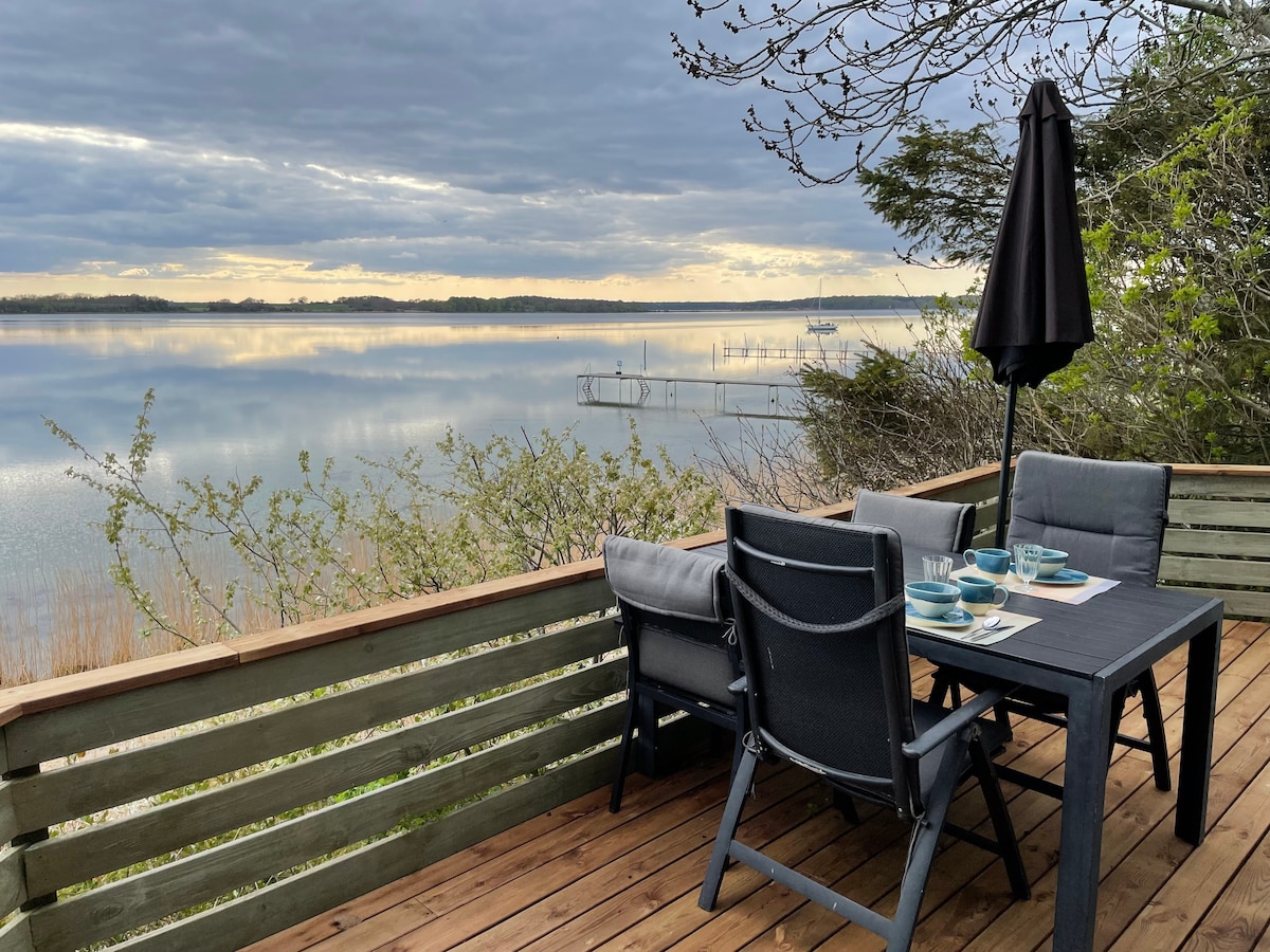 A wooden terrace is presented with a table and four chairs, overlooking calm waters. A black umbrella provides shade. The surrounding greenery and gentle waves contribute to a peaceful, nature-inspired setting.