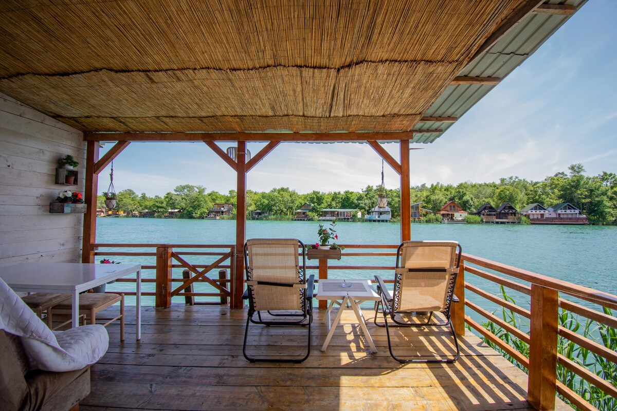 An inviting terrace is shown overlooking a tranquil river, featuring two lounge chairs and a small table. Wooden railings frame the space, complemented by a thatched roof providing shade. Lush greenery reflects along the water, with colorful cabins visible in the distance.