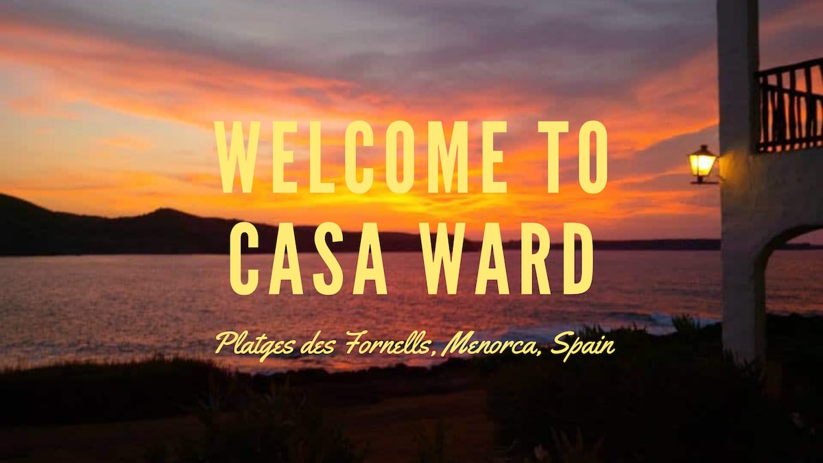 A vibrant sunset fills the sky with hues of orange, pink, and purple, overlooking the tranquil sea. A softly lit lantern on the wall adds warmth and ambiance to the view, while the surrounding landscape features lush greenery, welcoming guests to Casa Ward.