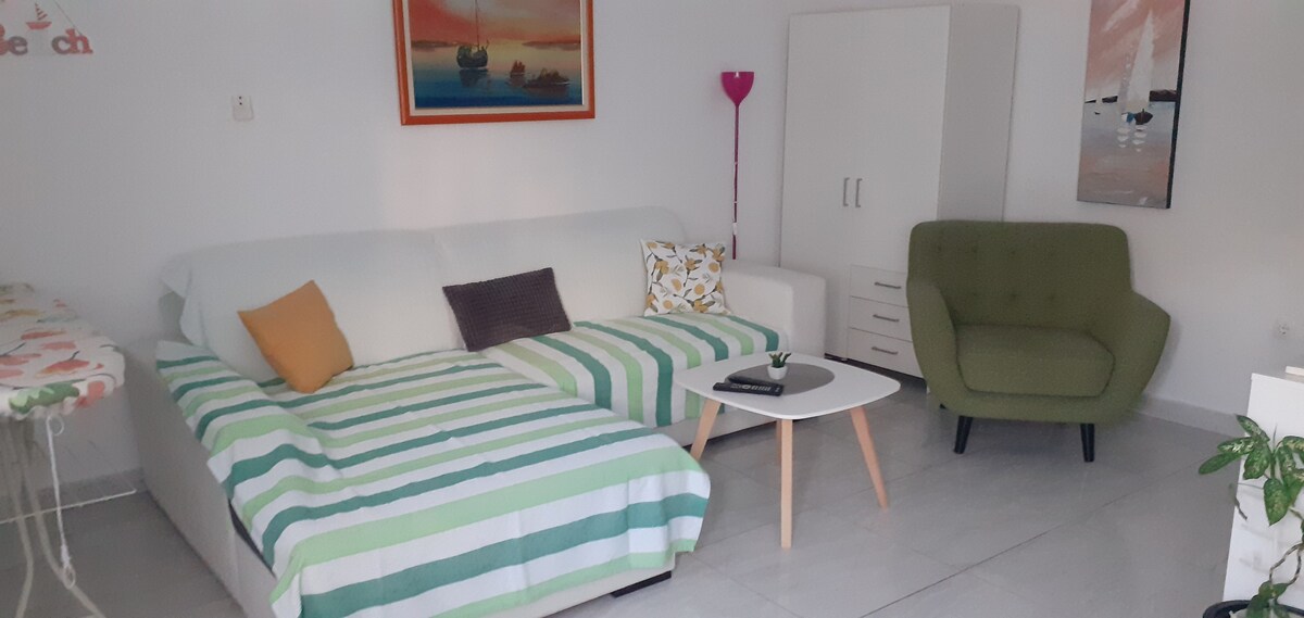 A cozy living area features a light-colored sofa with a striped blanket and colorful pillows. A green armchair sits nearby, accompanied by a small coffee table displaying a book. Light art pieces adorn the walls, complemented by a bright, inviting atmosphere.