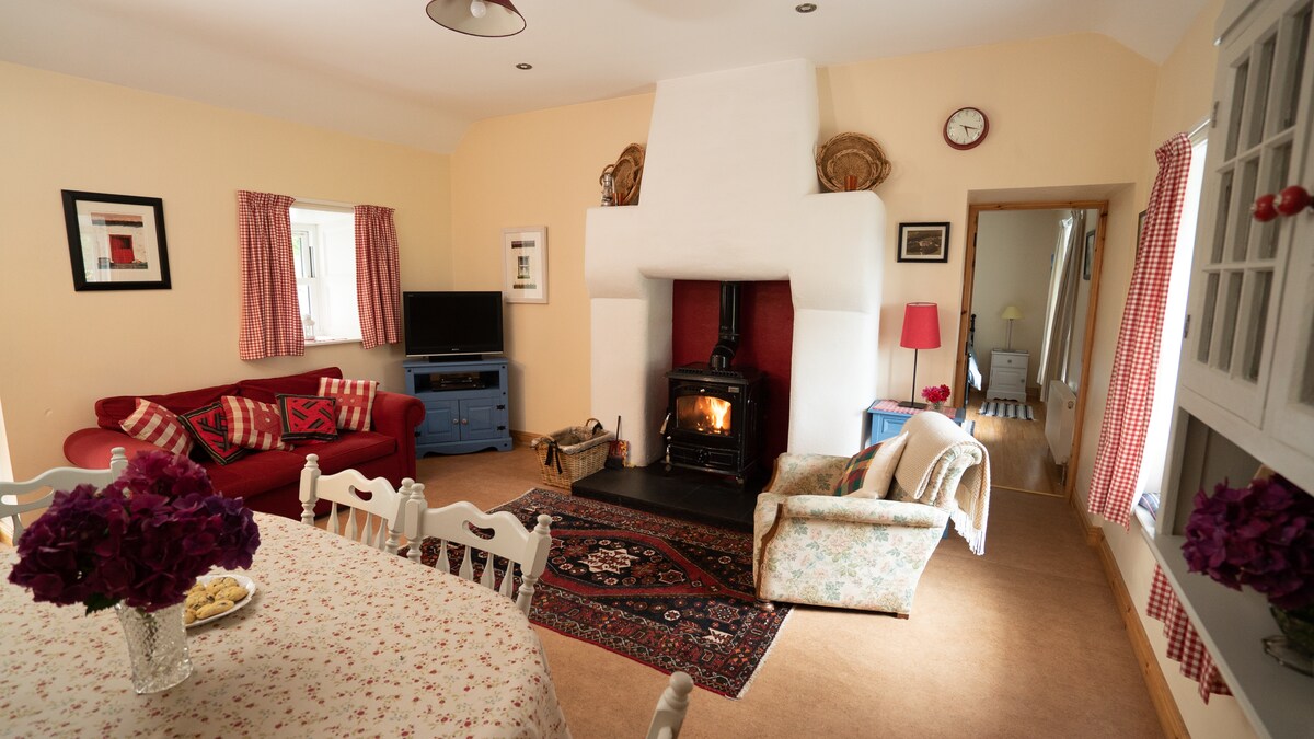 A living area is shown with a red sofa and armchair, arranged around a whitewashed fireplace with a fire burning. A dining table with floral tablecloth is set with chairs nearby. Light streams through the windows, complemented by soft curtains and wall art.