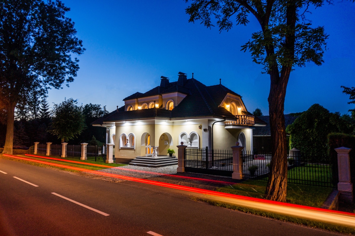 The villa is showcased at dusk, featuring elegant architecture with a well-lit façade. Its spacious exterior is framed by trees and a manicured garden, while the nearby road is illuminated by moving vehicle lights, creating a gentle contrast with the tranquil setting.
