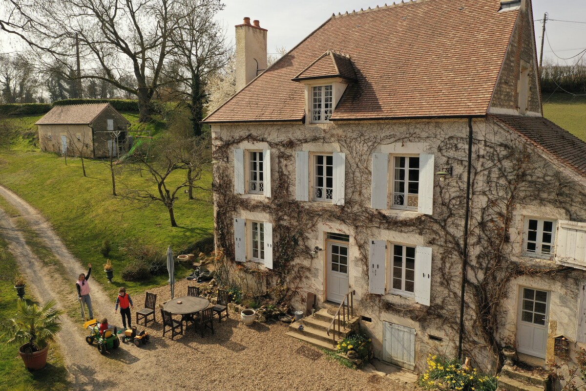 A traditional house with climbing vines covers its facade stands amidst a green landscape. At the front, a gravel area features a round table surrounded by chairs. Steps lead to the entrance, while a pathway winds through the property.
