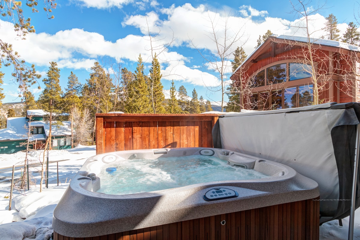 An outdoor hot tub is situated against a backdrop of snow-covered trees and mountains, with a wooden privacy fence surrounding it. Large windows of a nearby home reflect the blue sky and clouds, illuminating the space.