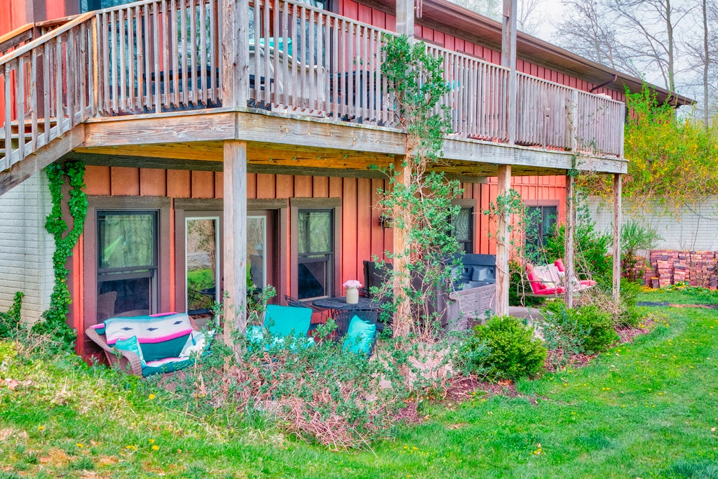 The exterior of a lower-level living space is framed by greenery and flowerbeds. A spacious patio area features comfortable outdoor seating, with several lounging chairs positioned near large windows. A balcony above enhances the outdoor ambiance, surrounded by natural elements and vibrant plant life.