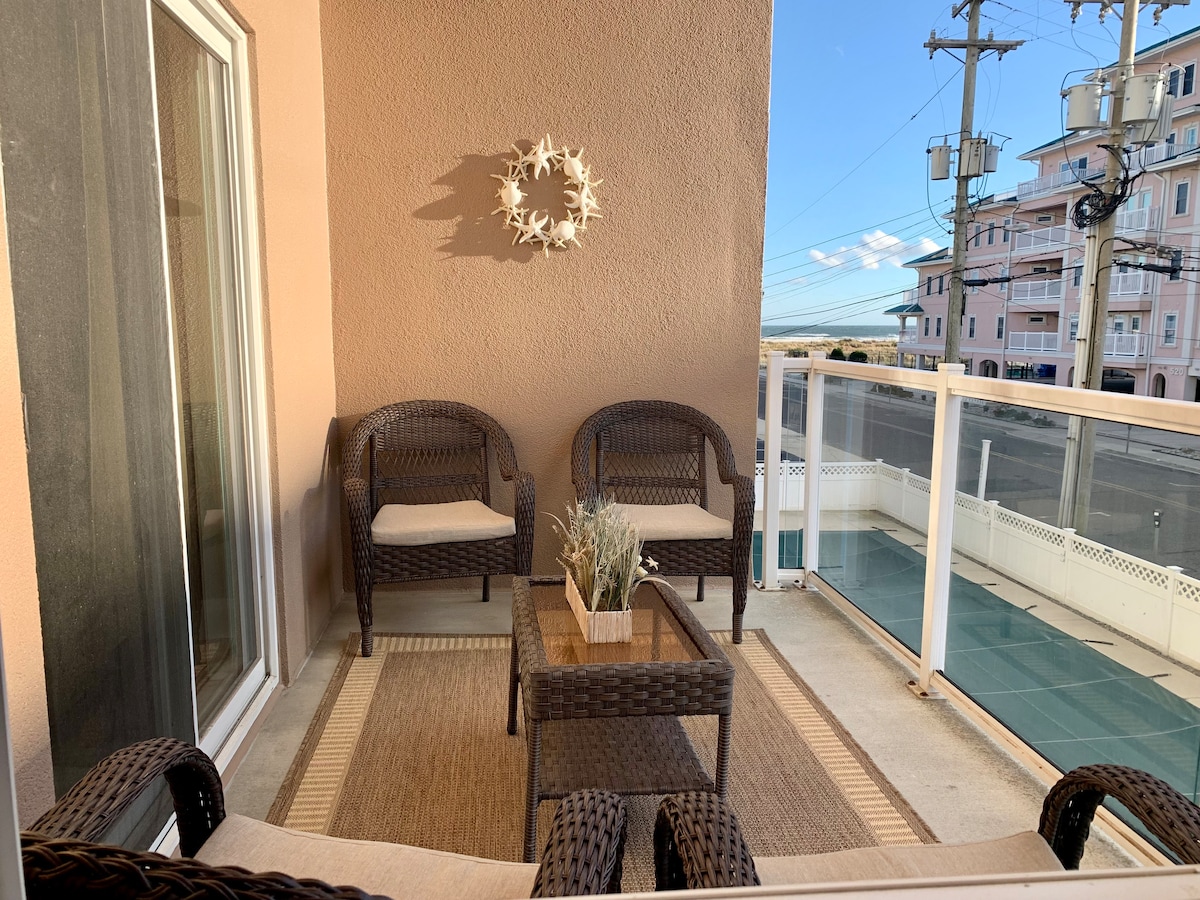 A private balcony features two woven chairs and a small table, all arranged over a neutral-toned rug. A decorative wreath adorns the wall, and the surrounding area includes views of nearby buildings and the sky. The space is designed for relaxation and enjoying the outdoor atmosphere.