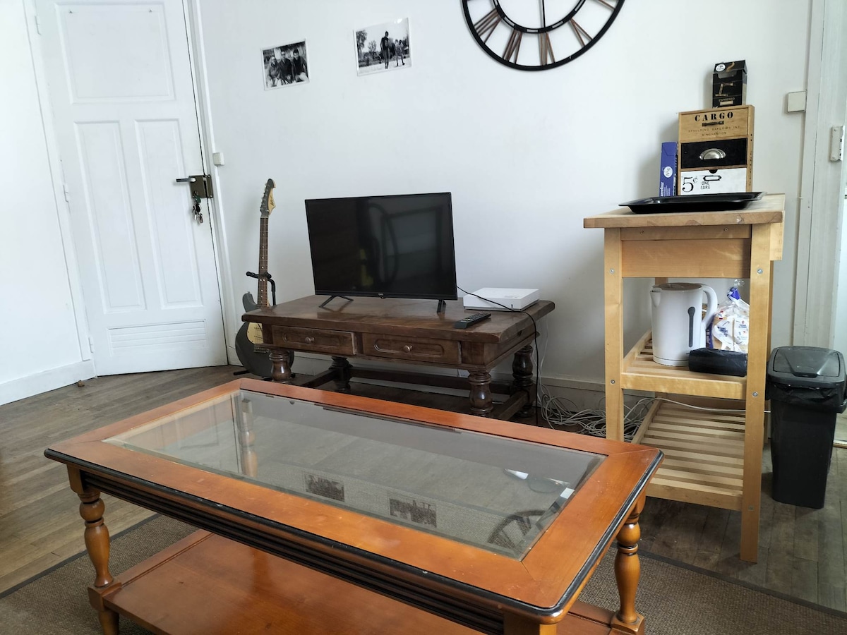 A cozy living area contains a vintage coffee table with a glass top, complemented by a television on a wooden stand. A guitar leans against the wall, and a small kitchen cart holds a kettle and a few kitchen essentials.