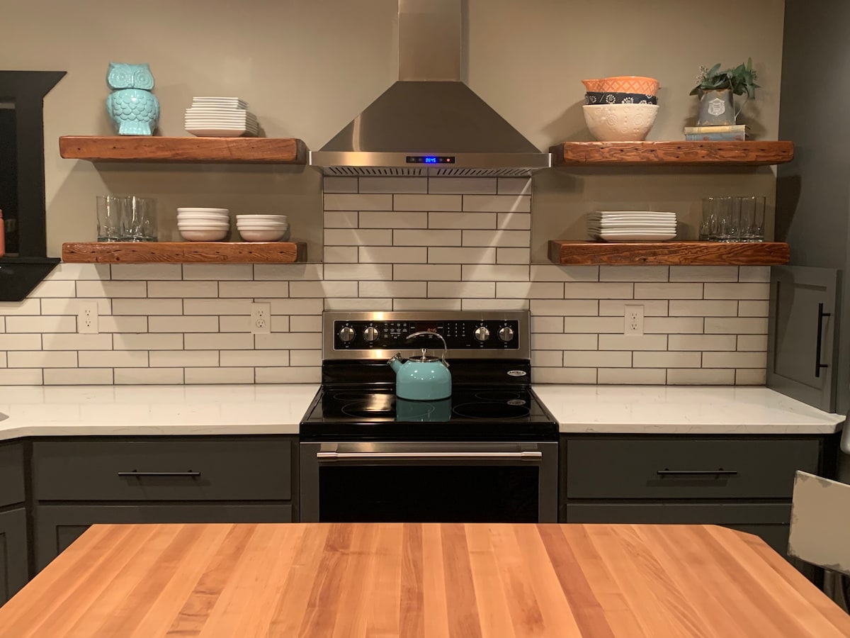 A modern kitchen area is highlighted by sleek countertops and a stainless-steel stove. Open wooden shelves display neatly arranged plates and glassware. A turquoise kettle adds a pop of color, creating an inviting cooking space. Subway tiles provide a clean backdrop.