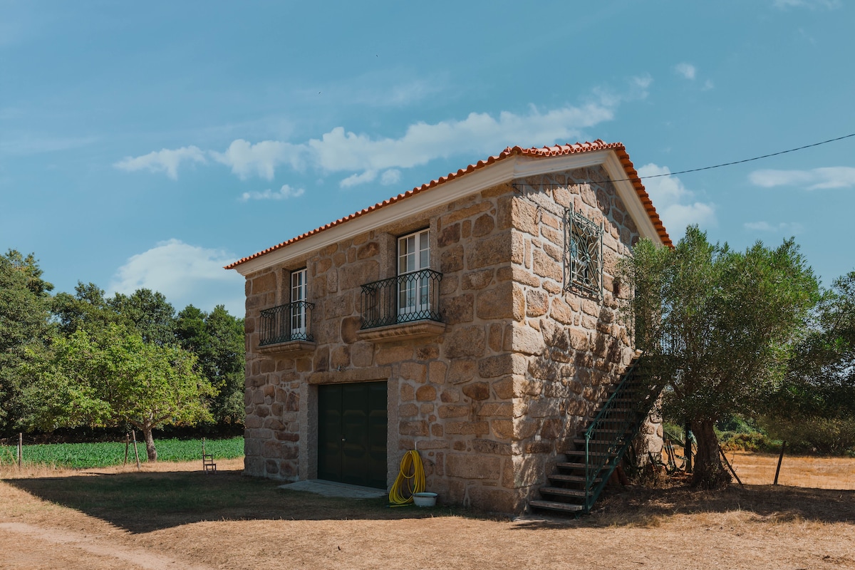 A rustic stone house with dual balconies overlooks a sunlit open field. An outdoor staircase leads to the upper level, surrounded by olive trees and greenery. The structure showcases classic architecture with a red-tiled roof and is set within a serene rural landscape.