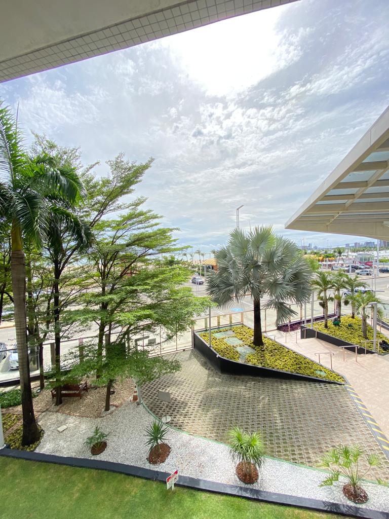 A garden area is visible, featuring a variety of green plants and palm trees. A paved pathway leads to the entrance, while the surrounding area includes traffic and city life. The sky is partly cloudy, allowing natural light to illuminate the scene.
