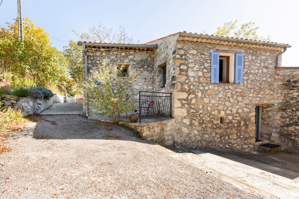 A charming stone house is positioned within a tranquil mountainous setting. The entrance features rustic stone walls and blue shutters. A small private terrace is visible, surrounded by greenery, with chairs placed invitingly. The pathway leading to the house is lined with gravel.