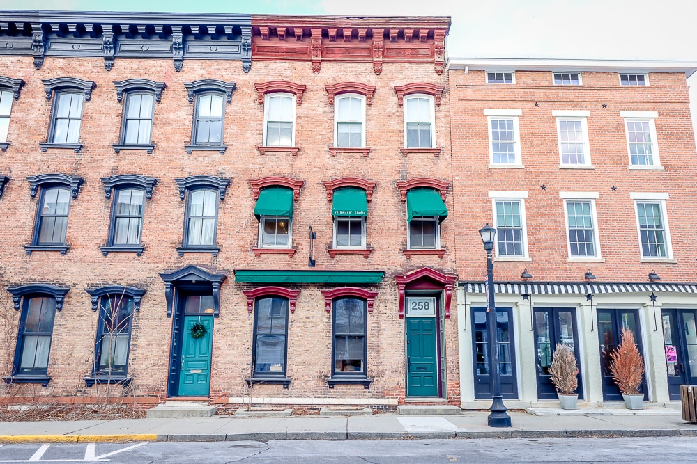 The exterior of a historic brick townhouse is displayed, featuring large windows with green awnings and a decorative door framed by brick. The surrounding architecture includes other brick buildings with varying styles, contributing to the charming streetscape.