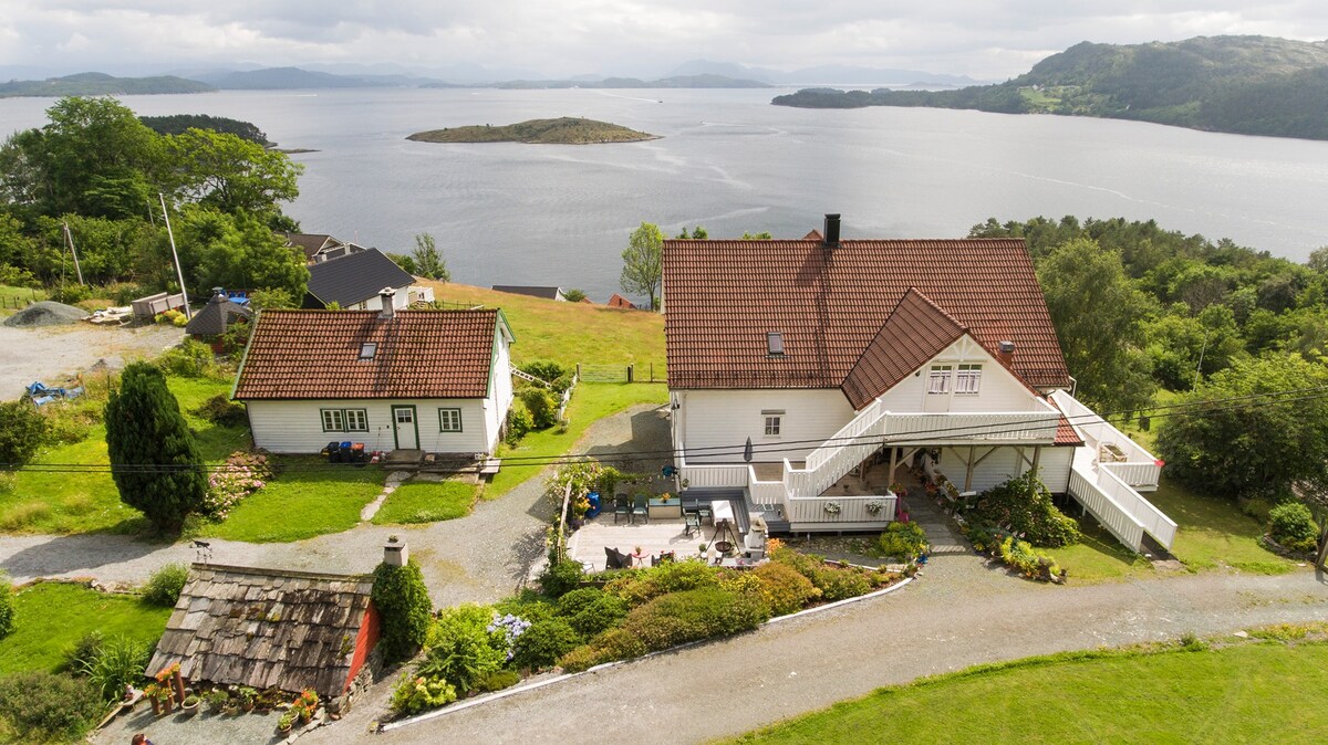 A scenic view of a property comprising two buildings, one with a steeply pitched roof and an outdoor staircase. Lush gardens surround the structures, and a lake with an island is visible in the background under a partly cloudy sky.