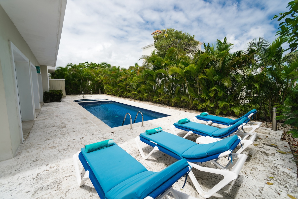 A private pool is framed by a smooth stone patio, surrounded by lush greenery. Several lounge chairs in vibrant blue line one side of the pool, offering a space for relaxation. The sky features soft clouds, creating a serene atmosphere.