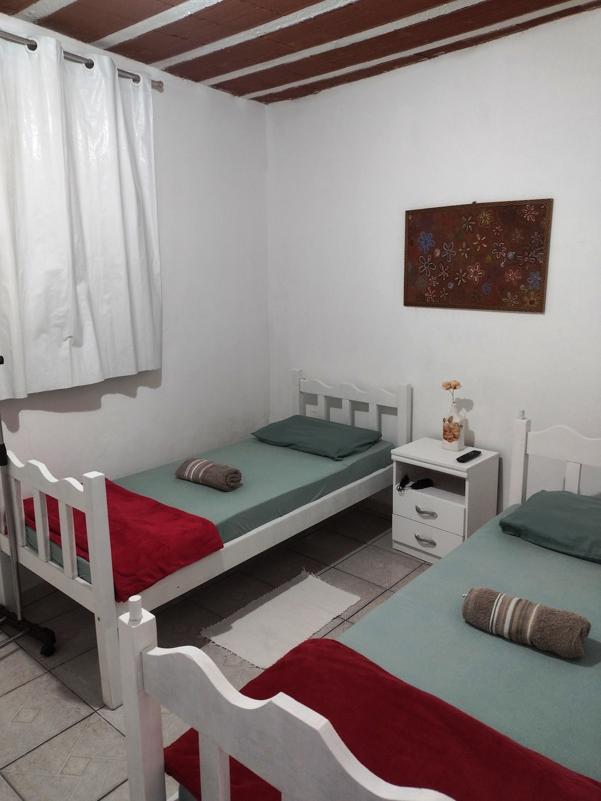 A room features two single beds draped in light green covers, each accompanied by a rolled towel. A white bedside table is positioned between the beds, topped with a small decorative item. Natural light enters through a window dressed with white curtains, illuminating the space.