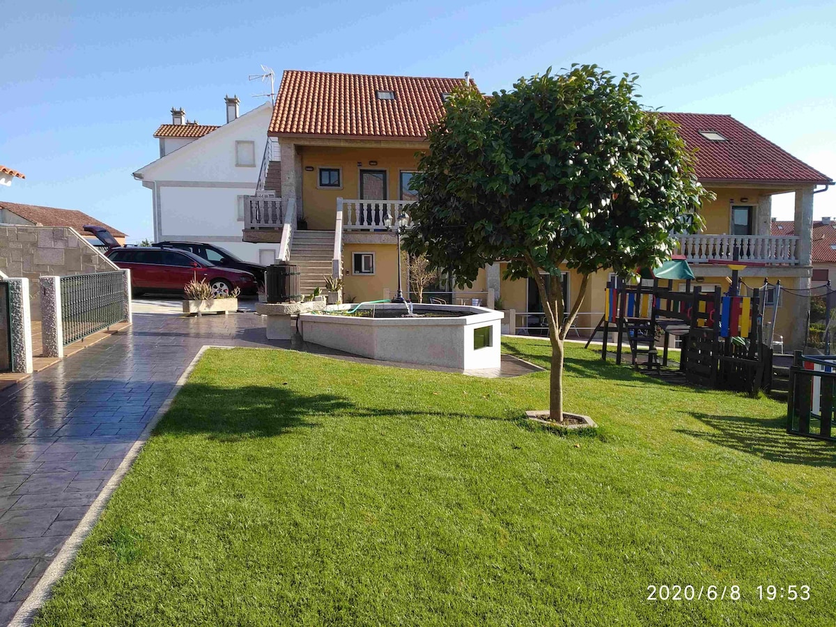 The image displays a well-maintained outdoor area featuring a lush green lawn, a fountain, and colorful playground equipment. Two apartment buildings are visible in the background, complemented by a clear blue sky. Parking is available nearby on a paved surface.