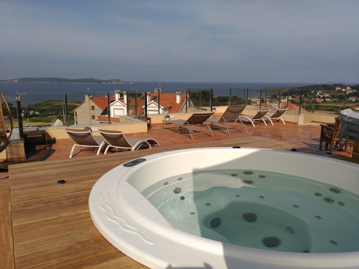 An outdoor terrace features a spacious Jacuzzi surrounded by loungers, offering views of the sea and surrounding landscape. The area is designed for relaxation, with wooden flooring and glass fencing ensuring an unobstructed vista.