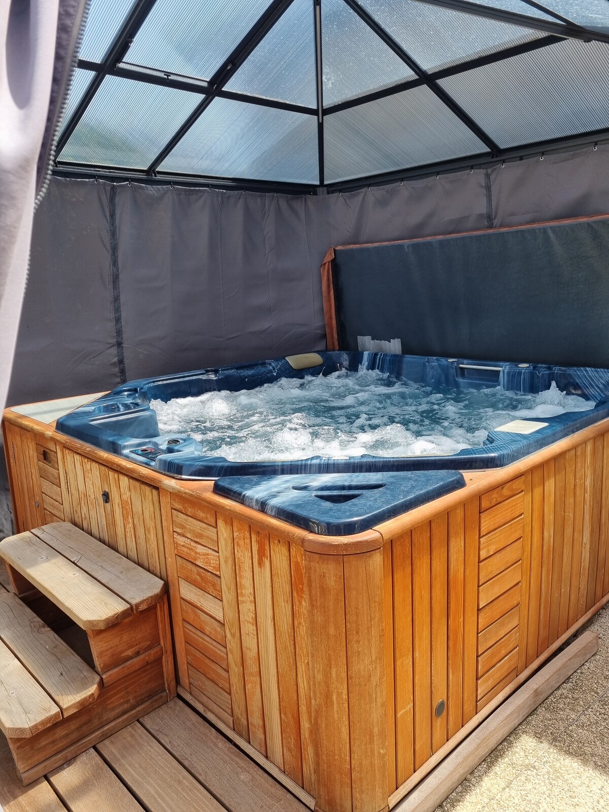 A private outdoor jacuzzi is enclosed within a protective canopy, featuring wooden paneling and steps for easy access. The bubbles create a relaxing atmosphere, inviting moments of leisure and comfort. Soft lighting enhances the serene setting surrounded by a natural landscape.