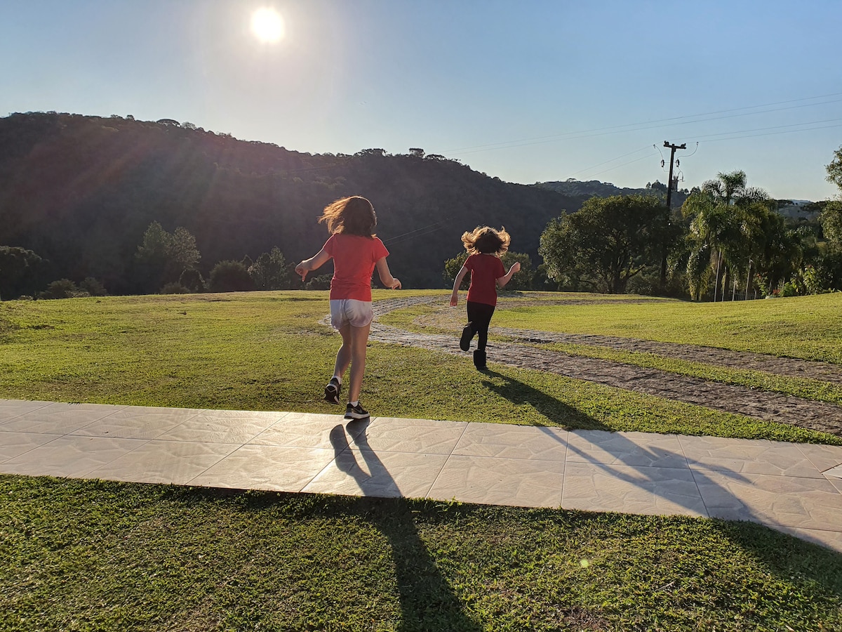 Two children are seen running on a stone path in a grassy area, with rolling hills and a bright sun illuminating the scene. The background features trees and a clear sky, creating an open and spacious atmosphere.
