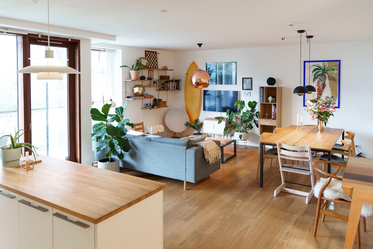 A spacious living area features a combination of a light wood dining table and a comfortable gray sofa adorned with cushions. Large windows allow natural light to fill the room, while a modern television is positioned on a wooden unit. Potted plants add greenery to the atmosphere, enhancing the overall brightness.
