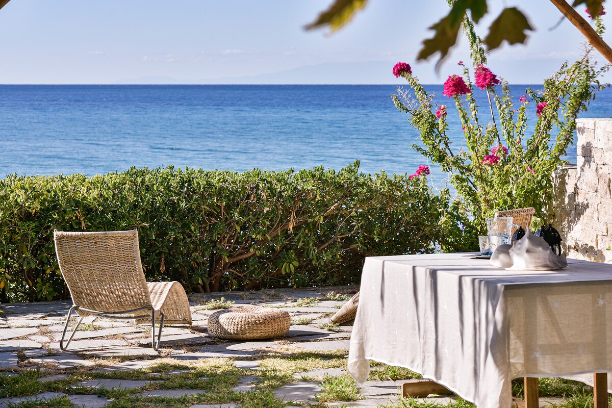 A serene outdoor space features a table set with decorative items and a comfortable chair facing the sea. Lush greenery and blooming flowers frame the scene, while the expansive ocean sparkles under clear skies in the background.