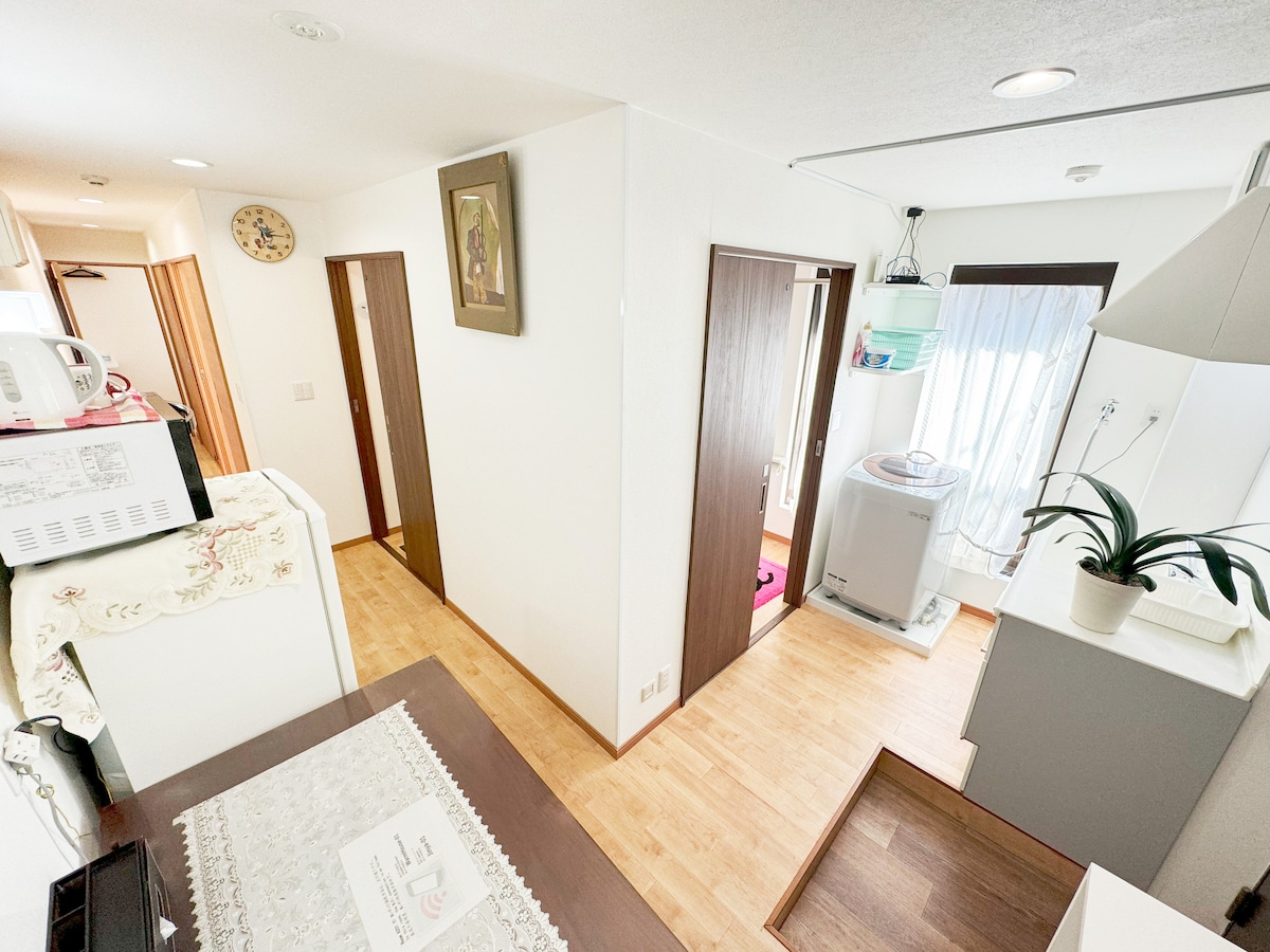 An open layout reveals a bright and airy space with light wood flooring. A doorway leads to a laundry area on the right, while a well-equipped kitchen is visible on the left. The room features soothing white walls and a window softly illuminating the interior.