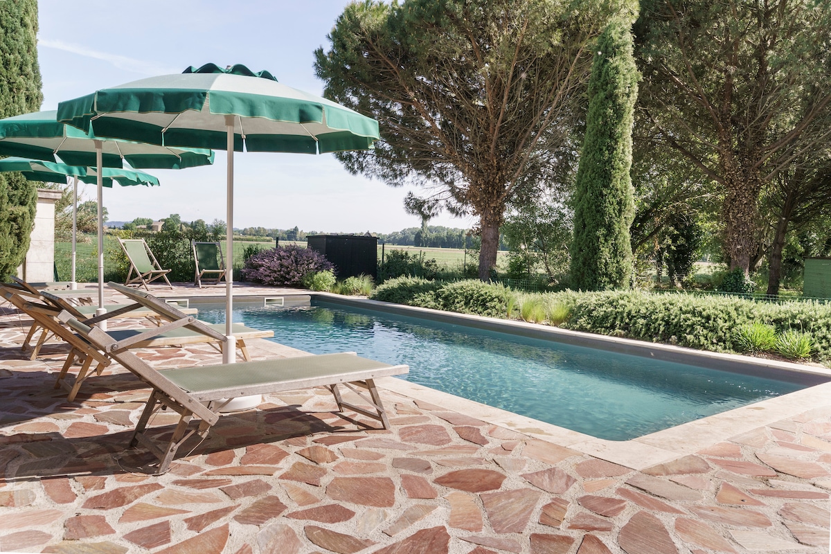 A serene pool area features wooden lounge chairs under green umbrellas, surrounded by lush greenery. The pool's water reflects the clear sky, creating a tranquil atmosphere. Stone patios border the pool, enhancing the outdoor space for relaxation.
