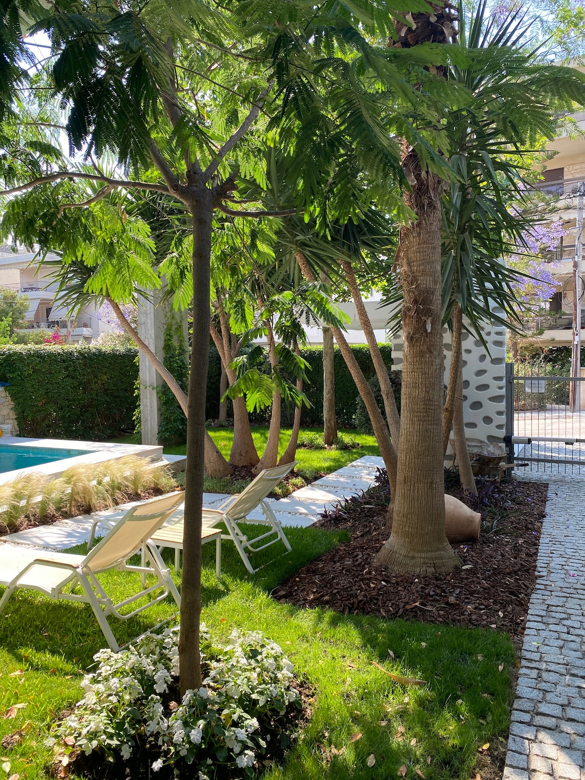 A sun-drenched garden area is framed by lush greenery, featuring tall palm trees and flowering plants. Lounge chairs are positioned on the grass near a refreshing pool, with decorative elements enhancing the tranquil setting. The space invites relaxation in a peaceful outdoor environment.