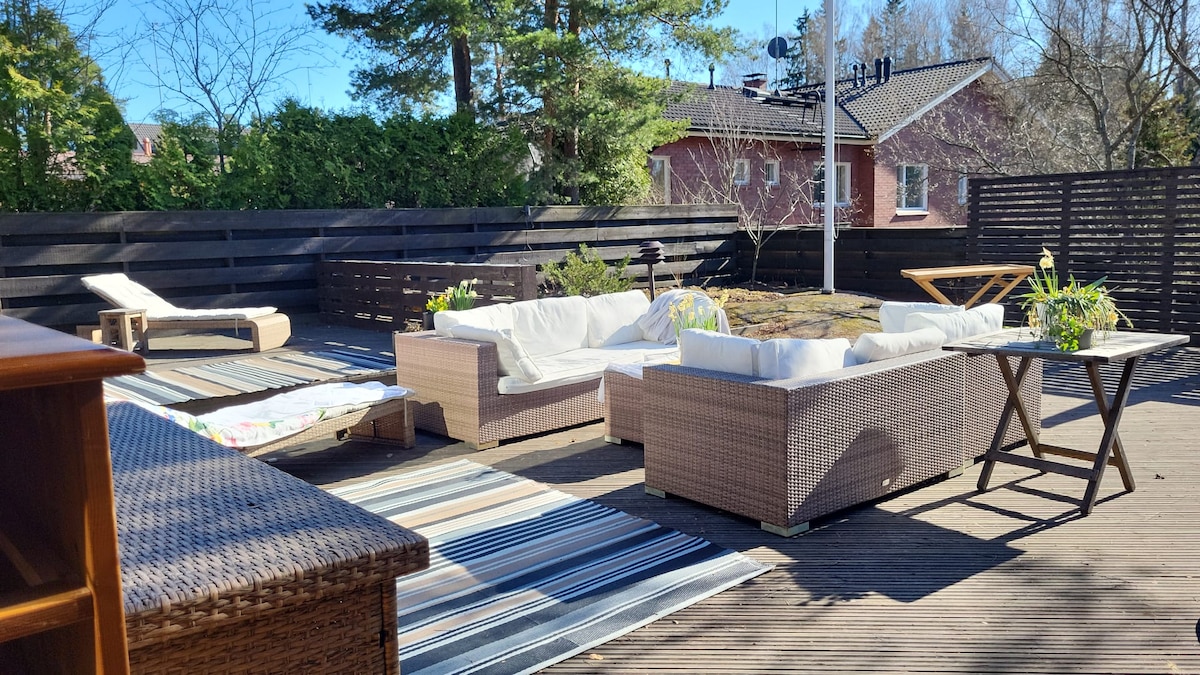An outdoor terrace is presented with comfortable seating arrangements, including several sofas and lounge chairs. Sunlight bathes the space, highlighting the wooden deck and soft area rugs. Planters with greenery add a touch of nature to the serene environment.