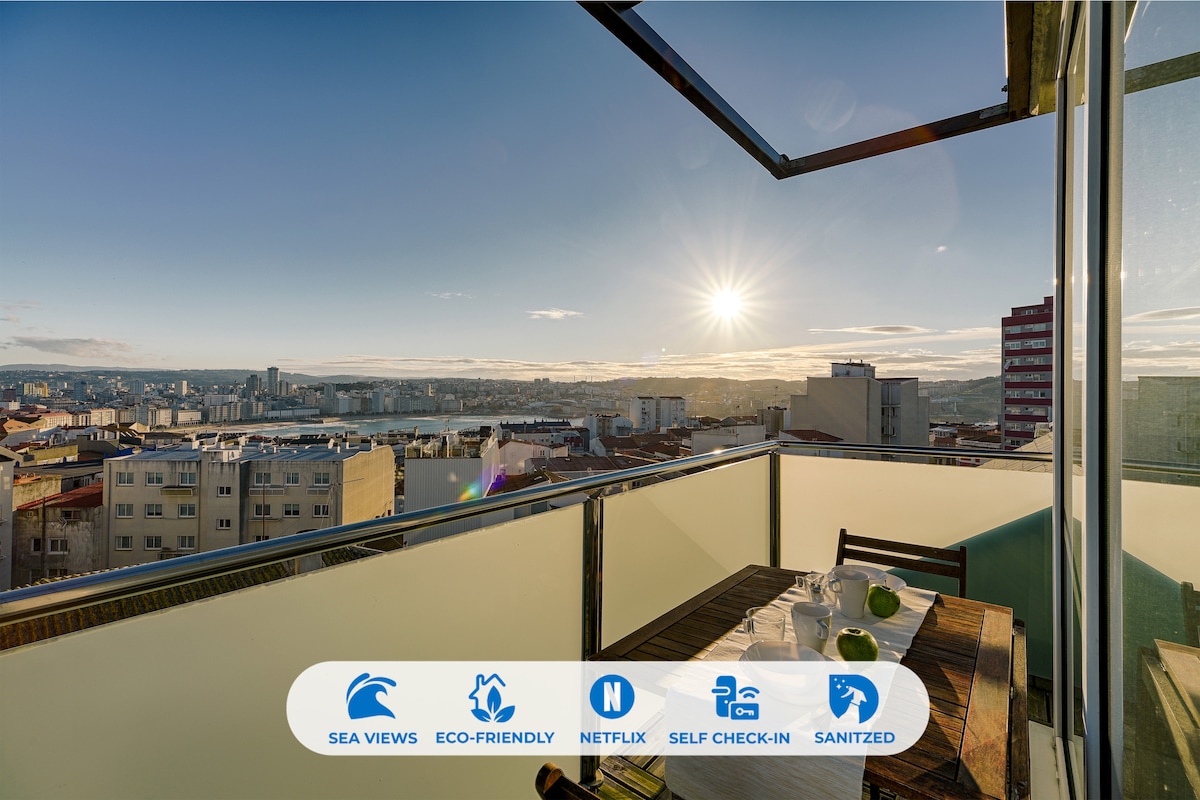 A terrace is shown with a dining table and chairs, offering panoramic views of the sea and the cityscape. The sunlight casts a warm glow over the space, enhancing the inviting atmosphere. The railing is glass, ensuring unobstructed views.