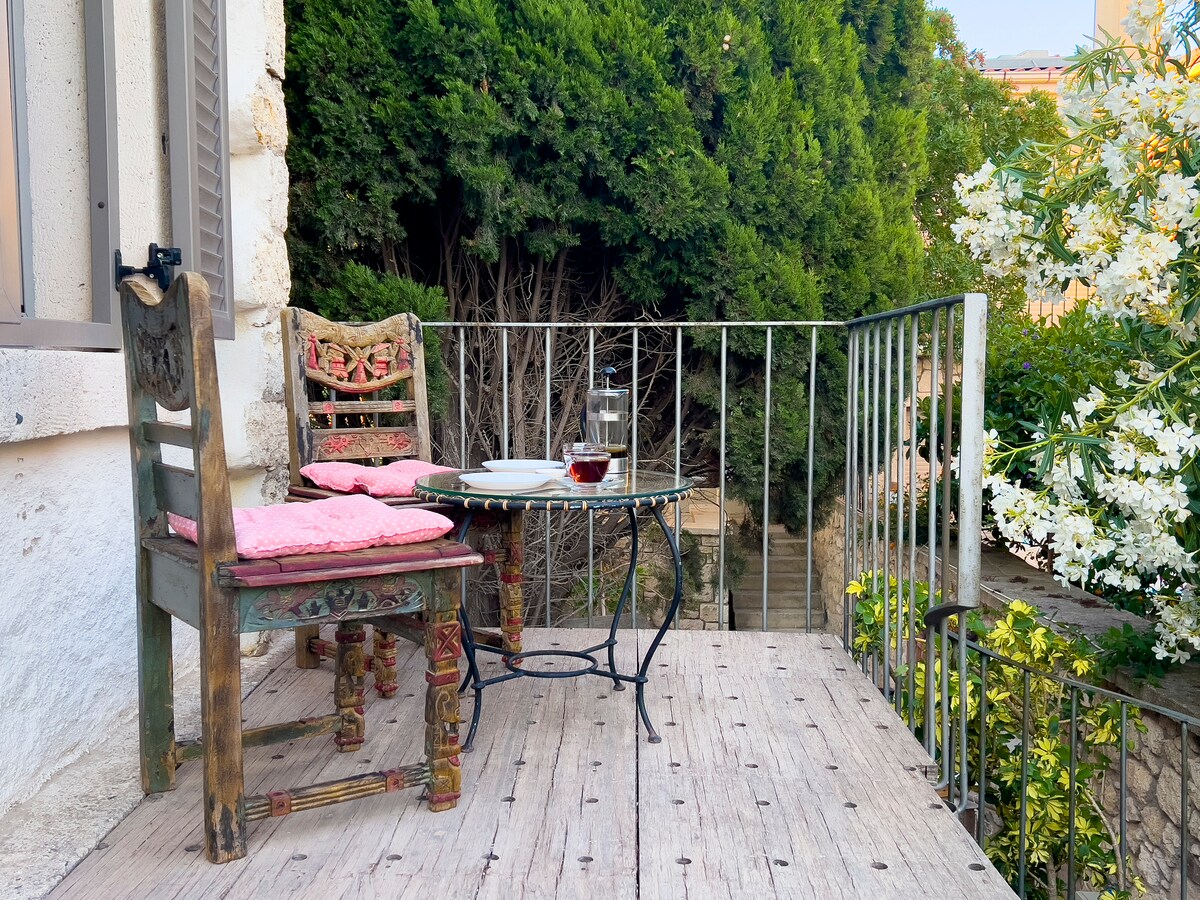 A small balcony area features two vintage chairs with cushions and a round table set with a plate and glass. Lush greenery and flowering plants surround the space, creating a serene outdoor retreat.