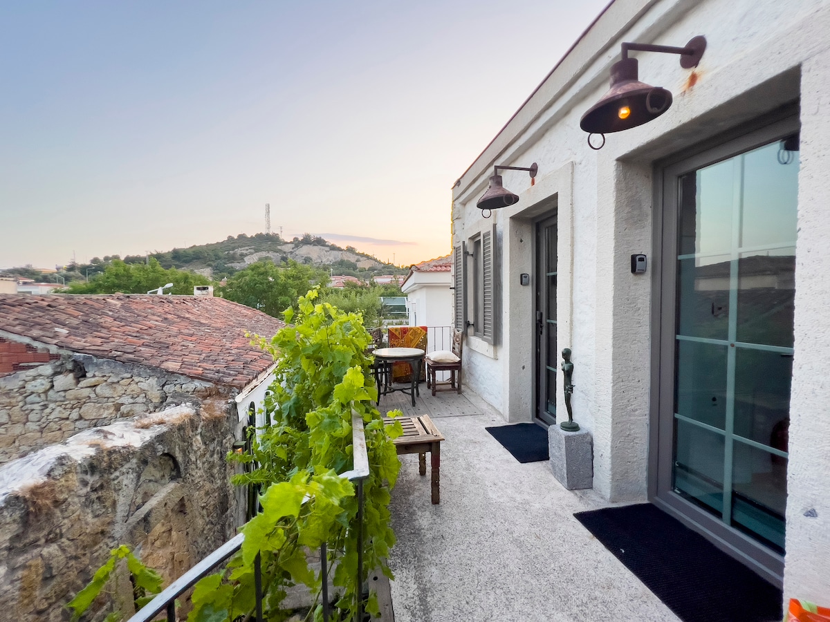 A serene balcony area is shown, featuring wooden seating and a table. Green vines climb the railing, while the backdrop includes a hillside silhouette under a soft evening light. Two doors lead into the interior, adding to the inviting outdoor space.