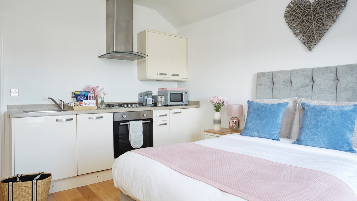 A cozy studio apartment features a double bed with blue accent pillows and a light pink throw. The kitchenette includes a gas hob, oven, and microwave, complemented by light cabinetry. Natural light fills the space, creating a welcoming atmosphere.