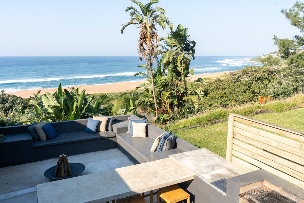 An outdoor terrace showcases comfortable seating arrangements set against a backdrop of lush greenery and the expansive ocean. A fire pit is positioned at the center, alongside a designated area for barbequing. Serene views of the beach and horizon contribute to the tranquil setting.