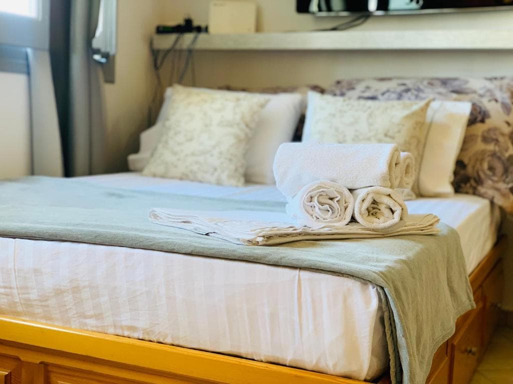 A spacious bed is neatly arranged with decorative pillows and a soft throw blanket. Fresh towels are rolled and placed on the bedding, contributing to the overall comfort of the sleeping area. A minimalist shelf is visible above the bed.