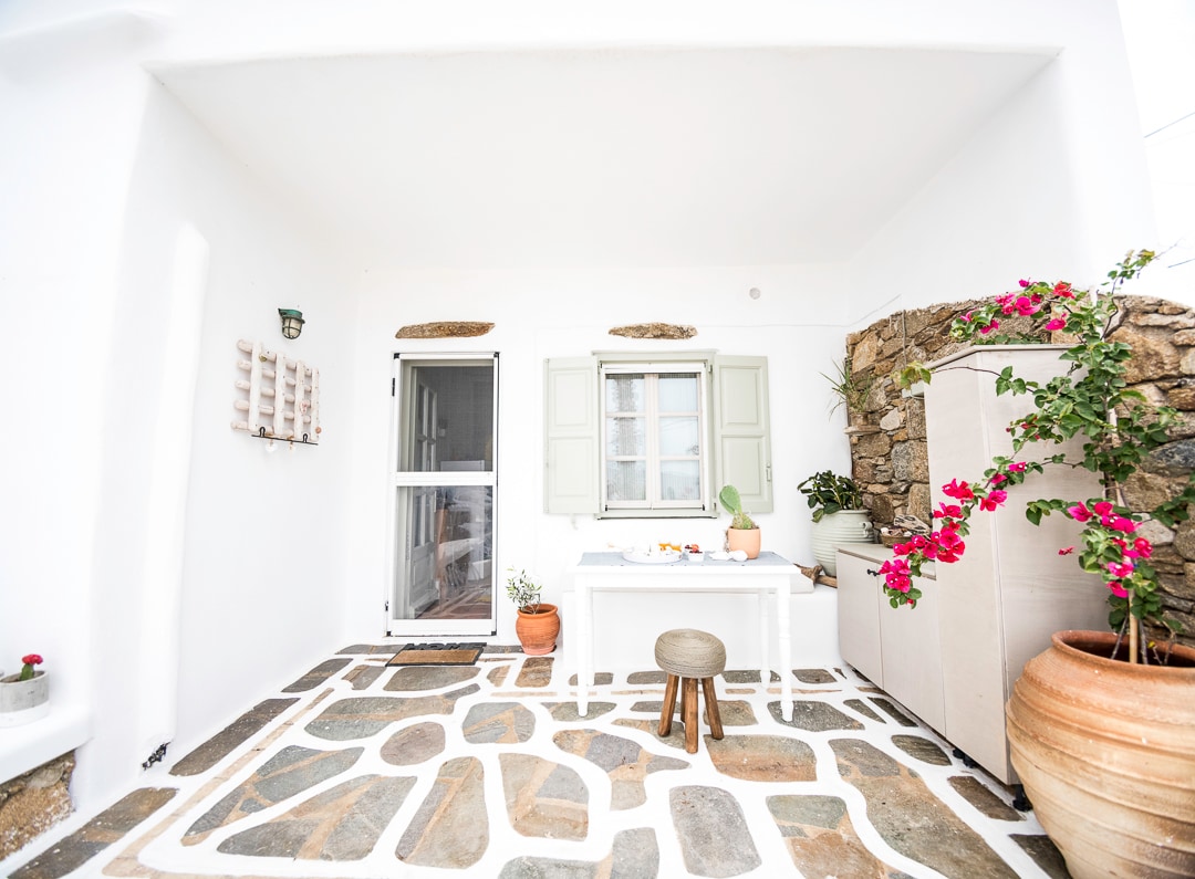 An inviting outdoor patio is presented with natural stone flooring and neutral white walls. A small dining table is surrounded by potted plants, while a decorative clay pot adds warmth to the space. Light-colored shutters frame the door and window, enhancing the serene ambiance.