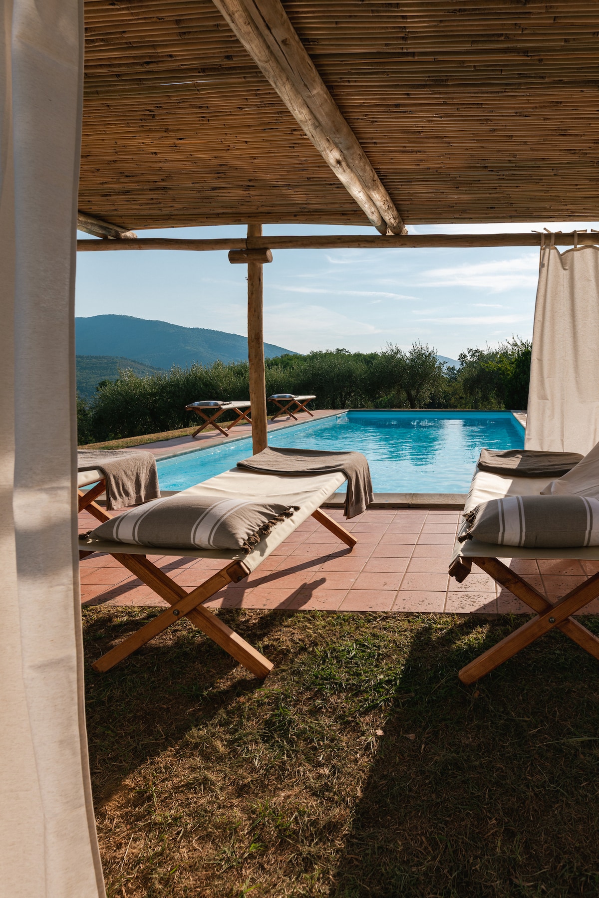 A serene view of a private pool set against a backdrop of rolling hills. Two sun loungers with soft, light-colored towels are positioned nearby, offering a relaxing spot to enjoy the surroundings. The area is shaded by a wooden structure, enhancing comfort.