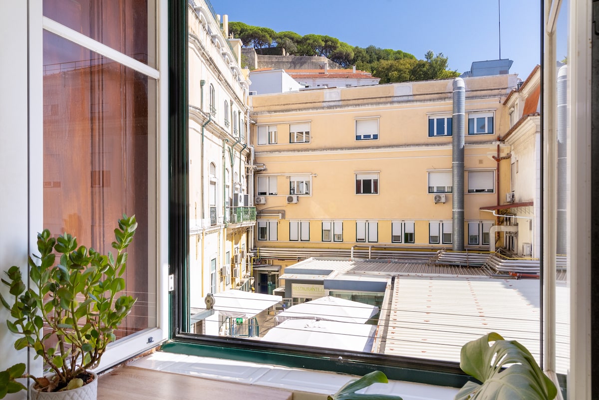 A view from a large window showcases a lively street with buildings in various shades of yellow and white. Potted plants are placed on the windowsill, adding a touch of greenery to the scene. Sunlight brightens the space, enhancing the inviting atmosphere.