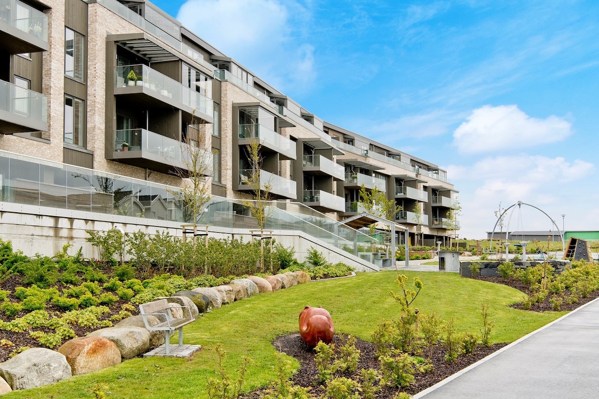 A modern apartment building with multiple balconies is positioned prominently in the image. Manicured green spaces feature a variety of shrubs and plants, along with a pathway leading through the landscaping. Benches are visible, providing seating areas amid the outdoor setting.