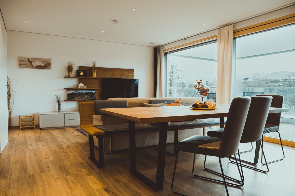A modern living area is presented, featuring a large dining table surrounded by four chairs. A comfortable sofa and entertainment unit are positioned against the wall. Large windows span the space, allowing ample natural light and offering views of the outside scenery.